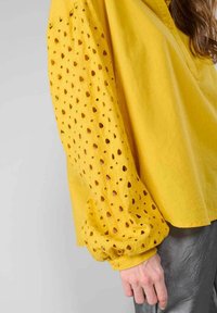 Blusa gialla a maniche lunghe con ritagli a occhio a forma di cuore, tessuto morbido e vestibilità comoda, abbinata a pantaloni grigi metallici.