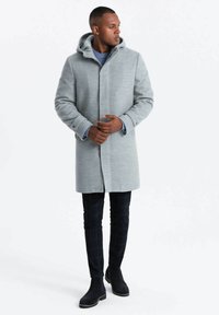 Abrigo de lana gris con capucha, frente de botones y bolsillos laterales. Usado sobre un suéter azul y combinado con pantalones de cuadros negros y botas oscuras.