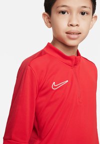 Haut de sport rouge Nike avec un col montant, fermeture à demi-zip, panneaux latéraux texturés et un logo blanc sur la poitrine.