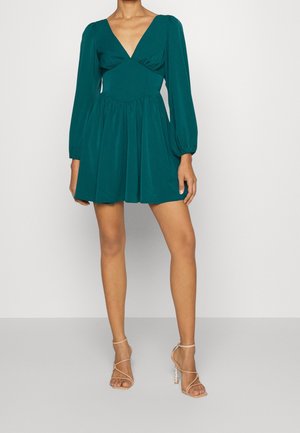 Kobieta ubrana w turkusową mini sukienkę z długim rękawem i głębokim dekoltem w kształcie litery V oraz beżowe, kuse sandały na wysokim obcasie, stojąca na jednolitym tle.