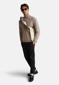 Mann trägt einen beigen Rollkragenpullover, schwarze Hose, schwarze Mokassins und eine Sonnenbrille, mit einer cremefarbenen und braunen Jacke über der Schulter.