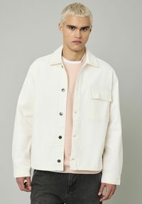 Un jeune homme aux cheveux blonds courts portant une veste bouton-up blanc cassé sur un sweat-shirt rose pâle et un jean gris foncé sur un fond gris.