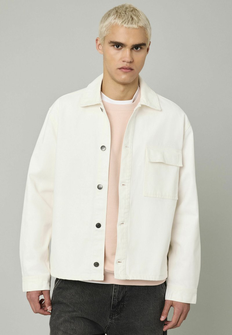 Un jeune homme aux cheveux blonds courts portant une veste bouton-up blanc cassé sur un sweat-shirt rose pâle et un jean gris foncé sur un fond gris.