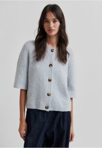 Cardigan tricotat de culoare gri deschis, cu mâneci scurte, având butoane mari, maro, pe față și o margine ribbed. Textură moale și croială lejeră.