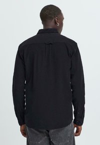 Black long-sleeve shirt made from textured fabric, featuring a standard collar and buttoned cuffs. Back has a hanger loop detail.