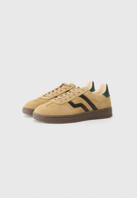 Tan suede sneakers with black stripes, brown rubber sole, and green heel accents. Features a textured finish and lace-up design.