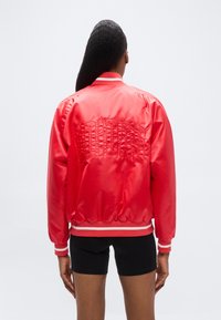 Άτομο με μακριά μαύρα μαλλιά ντυμένο με κόκκινο σατινέ bomber μπουφάν Adidas με ανάγλυφο λογότυπο στην πλάτη και μαύρο σορτς, στέκεται μπροστά σε λευκό φόντο.