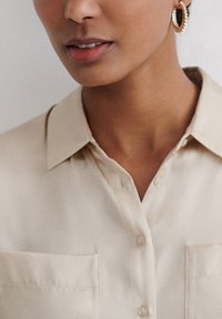 Beige Hemd mit Kragen, Knopfleiste, zwei Brusttaschen und glatter Textur; kombiniert mit goldenen Creolen.