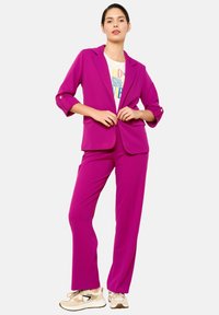 Completo fucsia con giacca su misura e pantaloni dritti. La giacca presenta maniche arrotolate e dettaglio con bottone singolo. Sotto, una t-shirt bianca con grafica.