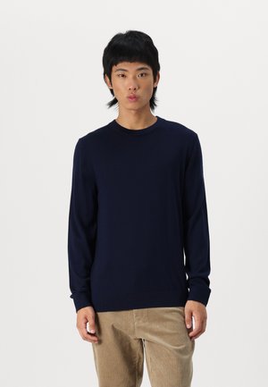 Maglione a maniche lunghe blu navy con scollatura a girocollo, texture liscia e polsini aderenti. Indossato con pantaloni di cotone a coste chiari.