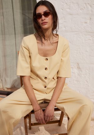 Vrouw draagt crème kleurige top met knopen en wijde broek, zittend op een houten stoel, met schildpadzonnebril en armband.