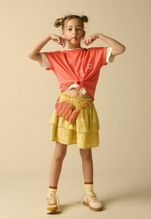 Jeune fille avec deux chignons portant un t-shirt corail noué, une jupe jaune, un sac en forme de poisson et des baskets, faisant une expression espiègle.