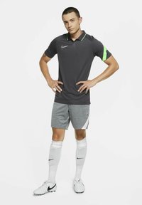 Graues Poloshirt mit grünem Akzent am linken Ärmel, kombiniert mit grauen Shorts und weißen Socken. Getragen mit weißen Fußballschuhen.