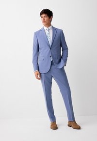 Helles blaues Sakko mit schlanker Passform, bestehend aus einem einreihigen Blazer und flachen Vorderhosen, kombiniert mit einem weißen Hemd und einer gemusterten Krawatte sowie braunen Schuhen.