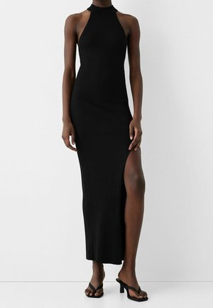 Abito lungo senza maniche nero a coste con scollatura halter e spacco alto fino alla coscia, indossato con sandali neri con tacco.
