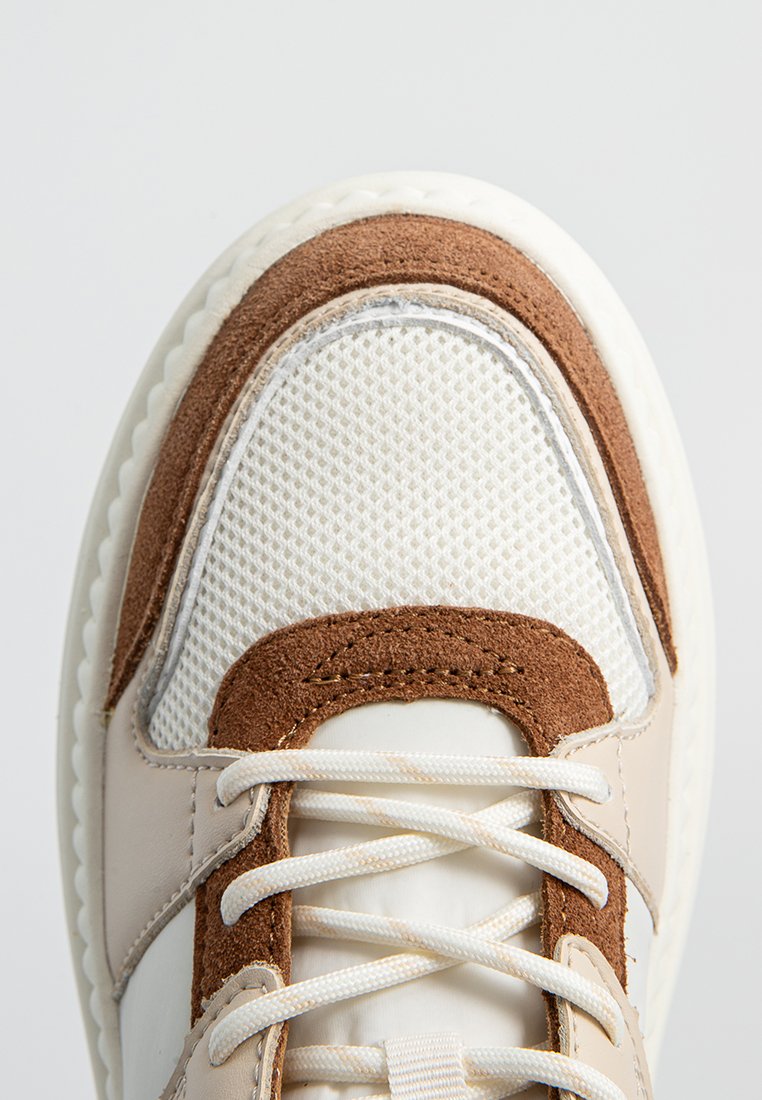 Sneaker con un design beige e marrone, caratterizzato da una cappuccio in rete, dettagli in camoscio e lacci bianchi. Parte superiore testurizzata e suola in gomma.