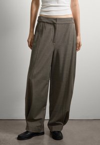 Persona vestida con pantalones marrones holgados, zapatos negros y una camiseta sin mangas blanca, de pie contra una pared gris claro liso.
