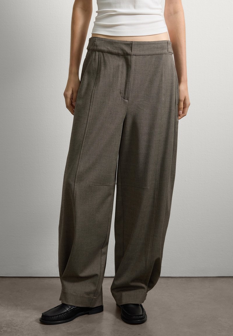 Personne portant un pantalon large marron, des chaussures noires et un haut sans manches blanc, se tenant devant un mur gris clair uni.