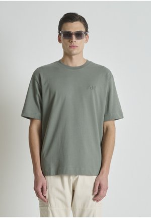 Man die een grijs oversized T-shirt, beige broek en donkere zonnebril draagt, staand tegen een effen lichte achtergrond.