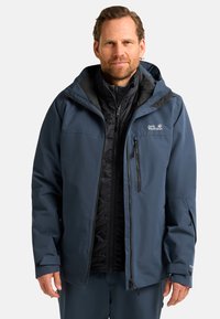 Giacca impermeabile blu navy con fodera isolante nera staccabile, tasche con zip, polsini regolabili e cappuccio. Caratterizzata da un logo sul petto.