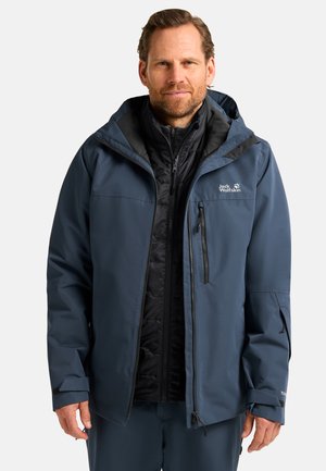 Marineblaues wasserdichtes Jacket mit abnehmbarer schwarzer isolierender Fütterung, Reißverschlusstaschen, verstellbaren Manschetten und Kapuze. Mit Logo auf der Brust.