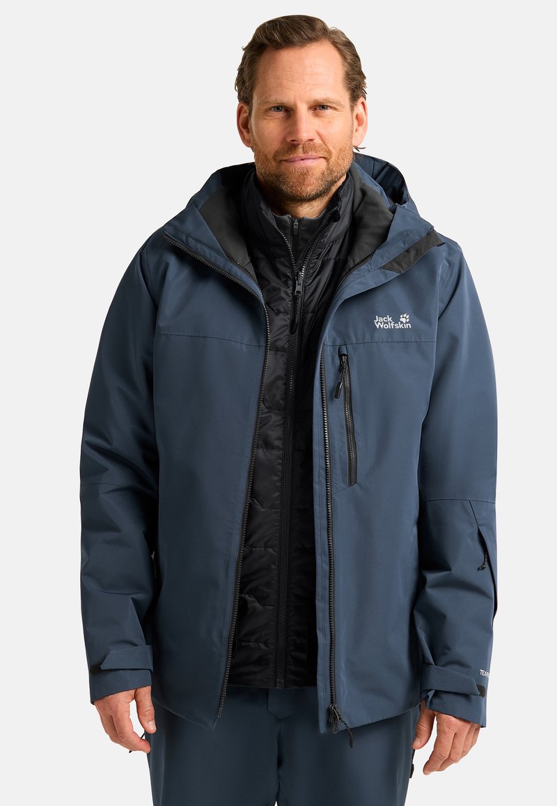 Giacca impermeabile blu navy con fodera isolante nera staccabile, tasche con zip, polsini regolabili e cappuccio. Caratterizzata da un logo sul petto.
