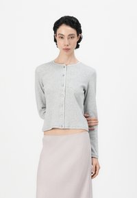 Grå knappet cardigan med rund hals, lange ermer og glatt tekstur. Brukt over et lys rosa skjørt.