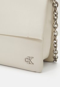 Krémová kožená kabelka s kovovým logem CK a stříbrným řetízkovým popruhem na bílém pozadí.