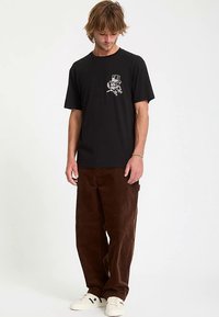 T-shirt nera in cotone con una stampa grafica sul petto, abbinata a pantaloni di velluto a coste marroni. Scarpe da ginnastica bianche completano l'outfit.