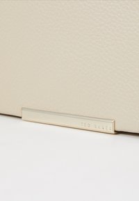 Beige nahkainen laukku, jossa on kultasävyinen metallikoriste, johon on kaiverrettu brändin nimi "TED BAKER". Suora, minimalistinen muotoilu.