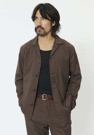 Barna gombos ing bemetszett gallérral, hosszú ujjakkal és foltzsebbel. Fekete atléta felsővel és barna övvel párosítva.