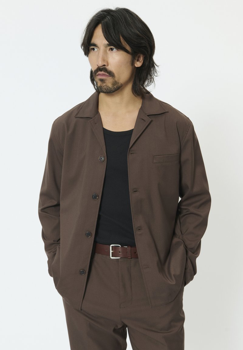 Brązowa koszula na guziki z wyciętym kołnierzem, długimi rękawami i naszywaną kieszenią. W połączeniu z czarnym topem na ramiączkach i brązowym paskiem.