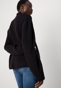 Strikket svart genser med høy krage, vide ermer og ribbestrikket tekstur. Kombinert med blå jeans, viser en behagelig passform og et enkelt design.