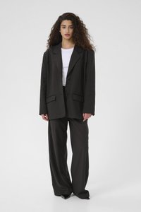 Černý oversize blazer se dvěma předními kapsami, kombinovaný s širokými kalhotami. Nositý přes bílé tričko, má hladkou texturu a zapínání na knoflíky.