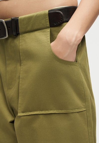 Χέρι μπαίνει στην μπροστινή τσέπη πράσινου χακί παντελονιού με μαύρη ζώνη, εστιασμένο στη μέση και την περιοχή του άνω μηρού.