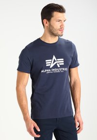 Tamsiai mėlynos medvilnės marškinėliai su balta grafika, logo ir tekstu "ALPHA INDUSTRIES", trumpomis rankovėmis ir apvali kaklo linija.