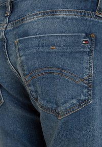 Vaqueros de mezclilla en un lavado medio, con un bolsillo trasero que tiene costuras naranjas y un pequeño parche de logo en rojo, blanco y azul.