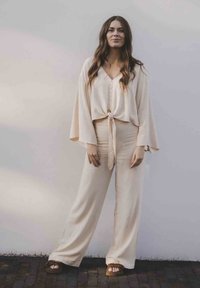 Beige tweed set met een geknoopte crop top met knopen en wijde broekspijpen. Zachte stof met een gladde textuur. Draag met bruine sandalen.