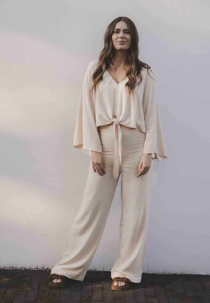Beige tweed set met een geknoopte crop top met knopen en wijde broekspijpen. Zachte stof met een gladde textuur. Draag met bruine sandalen.