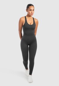 Zwart aansluitend jumpsuit met een ronde hals en brede banden, gemaakt van glad, rekbaar materiaal. Witte sneakers maken de look af.
