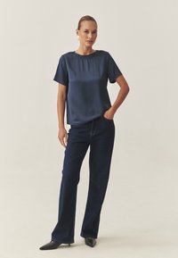 Navy Satinbluse mit kurzen Ärmeln, runder Halslinie; kombiniert mit dunklen Jeans, ausgestellten Jeans; spitze schwarze Schuhe. Glatte Textur, minimalistisches Design.
