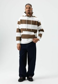 Sweatshirt met capuchon met horizontale bruine en witte strepen, voorzak en trekkoord. Gecombineerd met losse donkerblauwe jeans en zwarte schoenen.