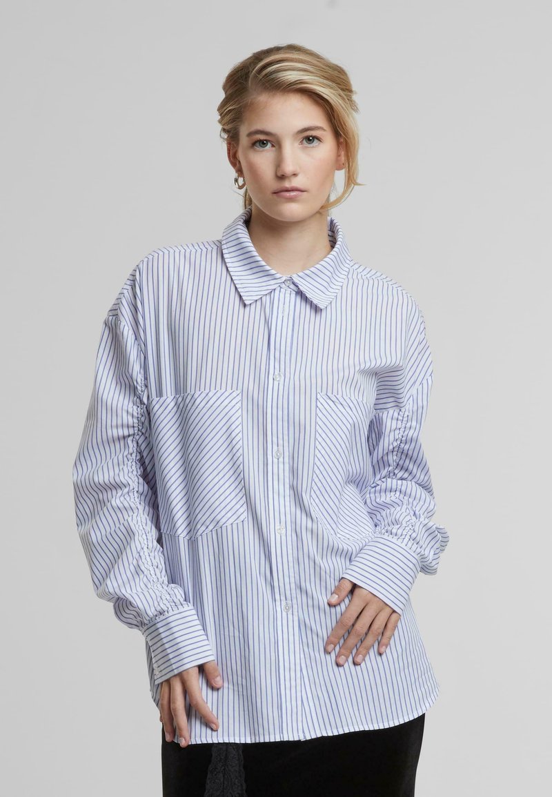 Camicia a righe azzurro chiaro e bianca, con colletto, vestibilità oversize, maniche gonfie e tasche applicate. Tessuto dalla texture liscia.