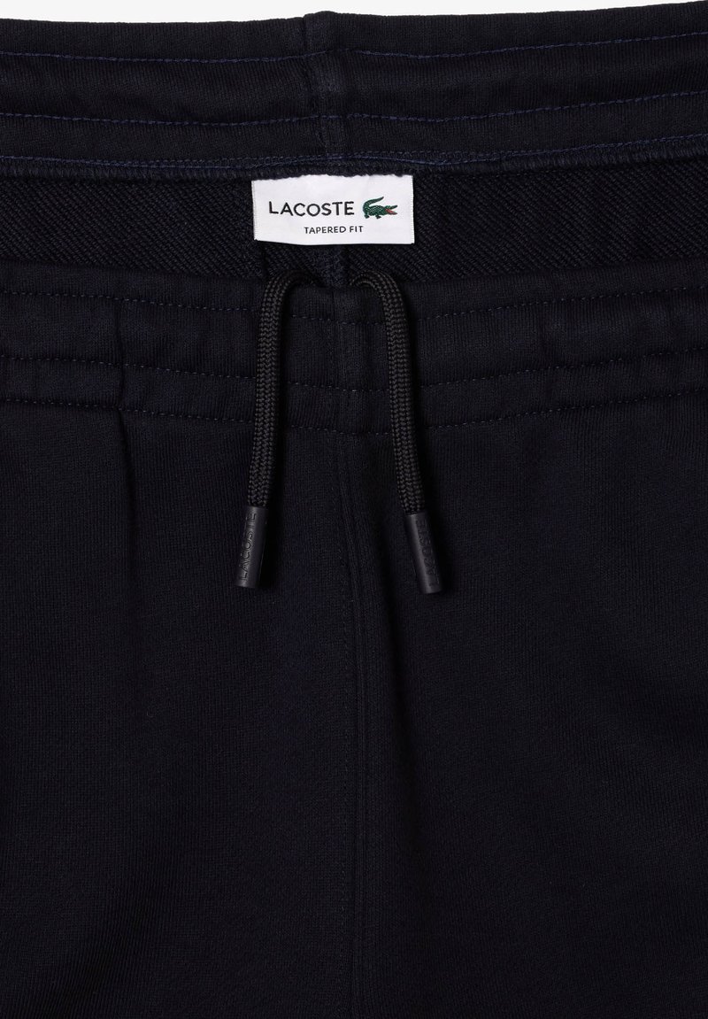 Pantalons de jogging noirs ajustés avec taille élastique, cordons de serrage noirs et étiquette blanche Lacoste avec logo crocodile vert.