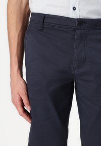 Pantalon bleu marine en tissu texturé, avec une fermeture à bouton à l'avant, des passants de ceinture et des poches latérales. Le design est décontracté et ajusté.