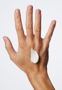 Vit kräm ligger på baksidan av en ljusbrun hand med utsträckta fingrar. Huden ser slät ut och bakgrunden är enfärgad vit.