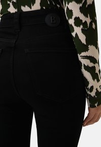 Osoba v černých těsně padnoucích džínech s vysokým pasem a dlouhým rukávem se zeleným a bílým vzorem, s rukou uvolněně položenou vedle sebe.