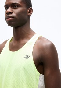Homme athlétique portant un débardeur de sport sans manches perforé New Balance jaune clair, légèrement tourné vers la gauche sur un fond blanc uni.