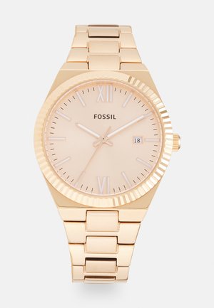 Fossil gouden-horloge met een gladde, ronde kast, minimalistische perzikkleurige wijzerplaat, Romeinse cijfers en een gekoppelde metalen armband. Datumaanduiding op 3 uur.
