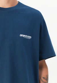Navy blue cotton T-shirt with a round neckline. Features a small white printed logo on the left chest area. Short sleeves, smooth texture.